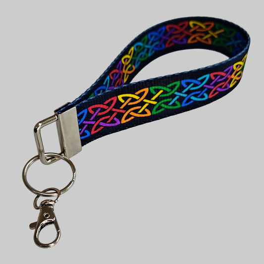 A fabric wristlet keychain strap with a vibrant rainbow-colored Celtic knot pattern on a black background, featuring a silver metal clasp and key ring on a light gray background.