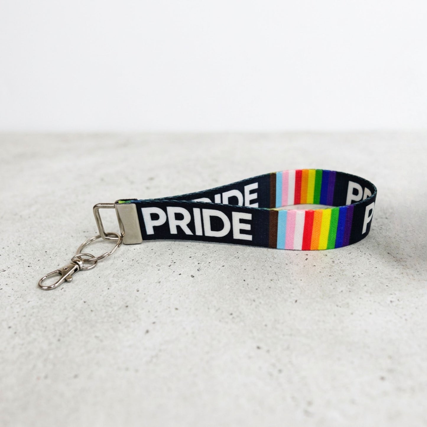 A fabric wristlet keychain strap with 'PRIDE' printed in bold white letters, featuring a black background and a vertical rainbow stripe pattern, finished with a silver lobster clasp and split ring, resting on a light gray textured surface.