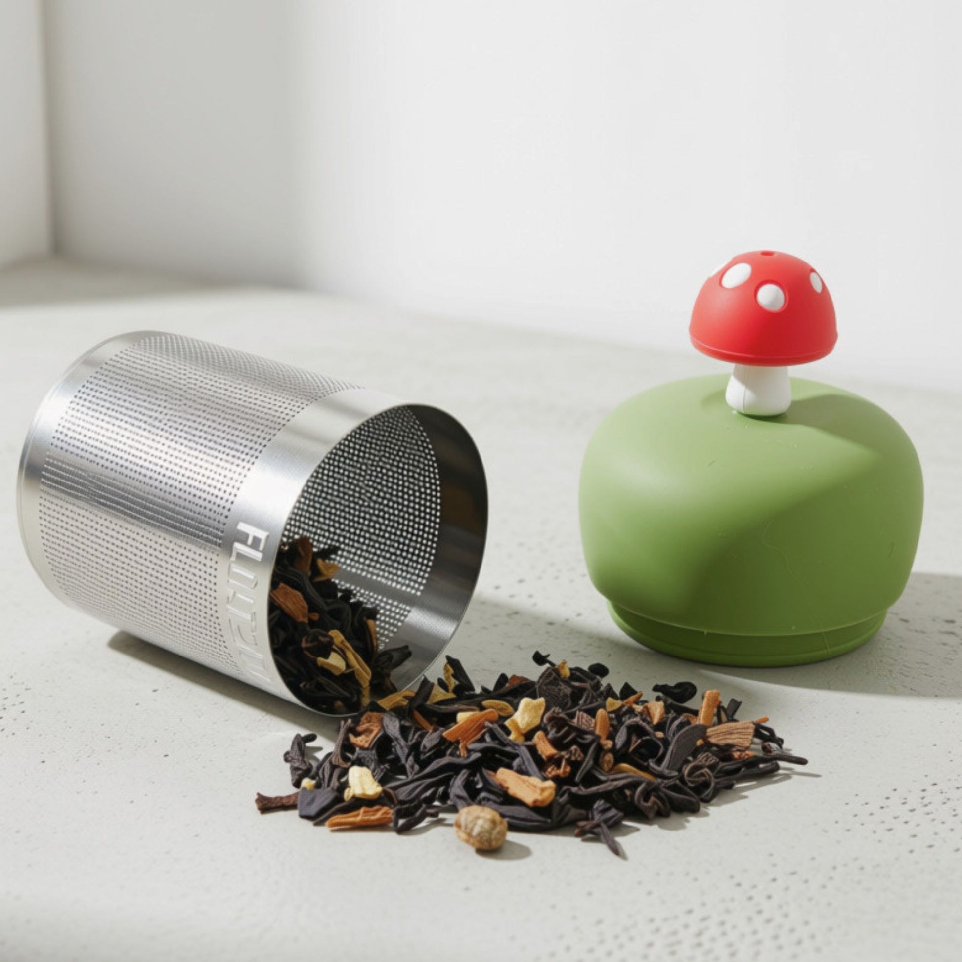 A stainless steel mesh tea infuser container lying on its side with loose tea spilled out, next to a green lid with a red mushroom topper on a light surface.