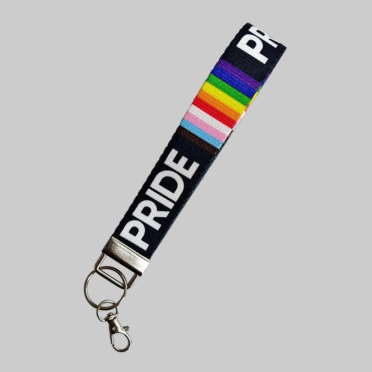 A fabric keychain strap with a black background, white 'PRIDE' text, a rainbow stripe section, and a silver metal clasp on a light gray background.